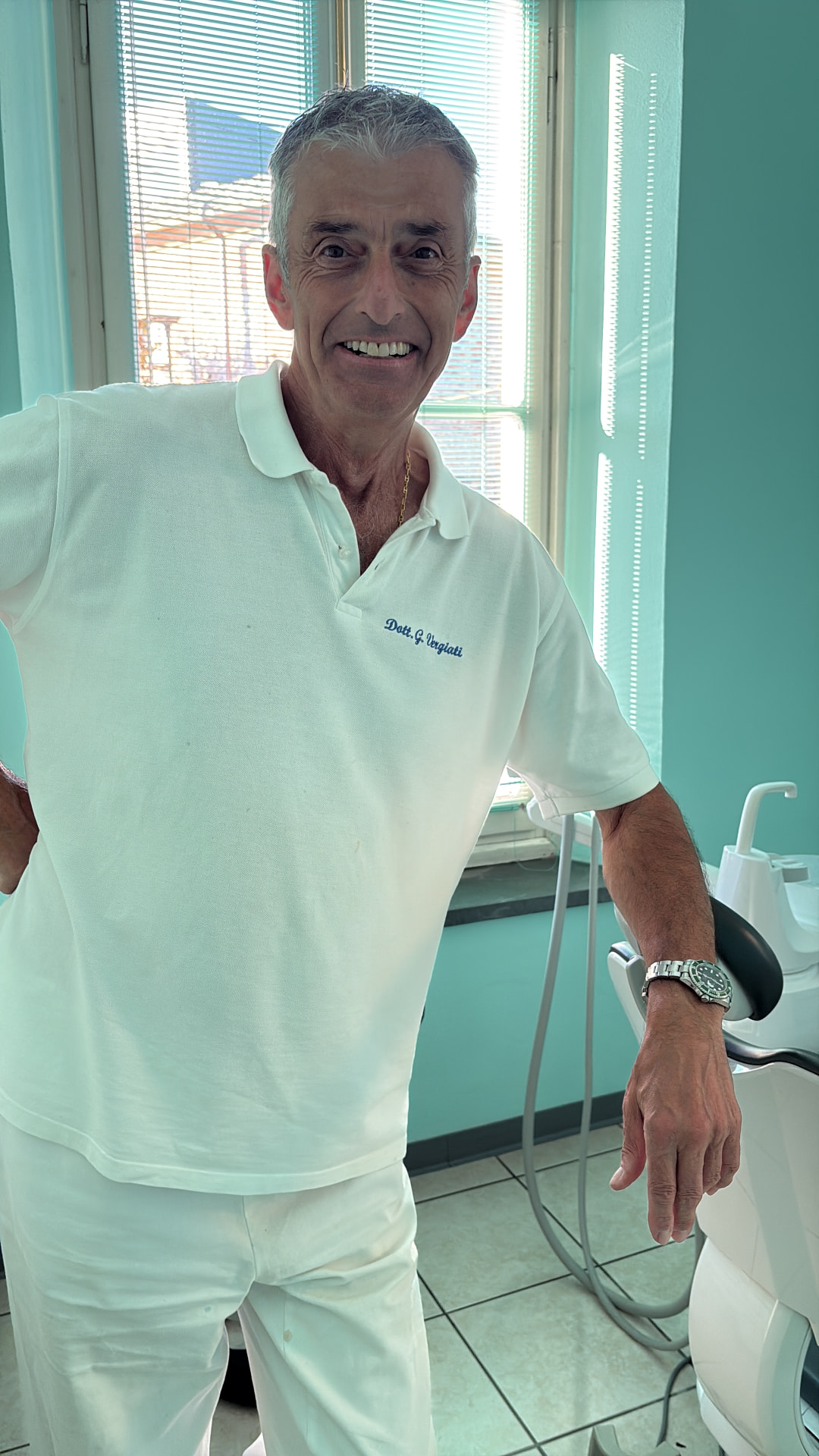 Il Dott. Giovanni Vergiati sorridente nello studio dentistico, in polo bianco ricamato, accanto alla poltrona odontoiatrica